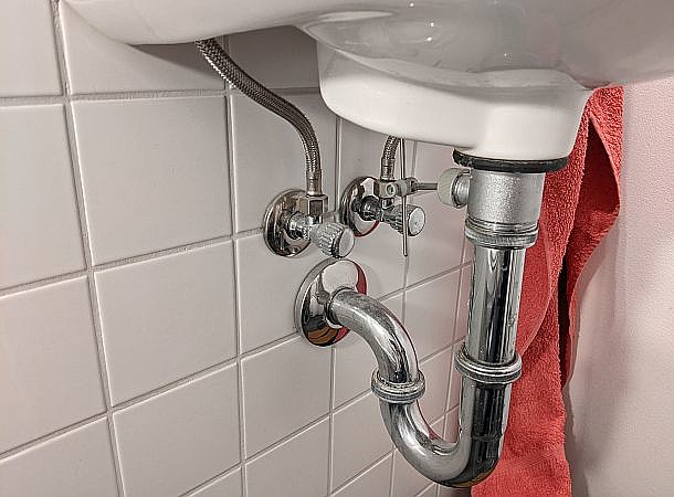 Blick unters Waschbecken: Ventil für Warmwasser, Kaltwasser, Abflussrohr, im Hintergrund ein rotes Handtuch