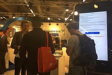Messestand mit co2online-Referentin und zwei Gästen, rechts das rund zwei Meter große Maxi-Smartphone mit der Handwerker-App Heizungsoptimierung